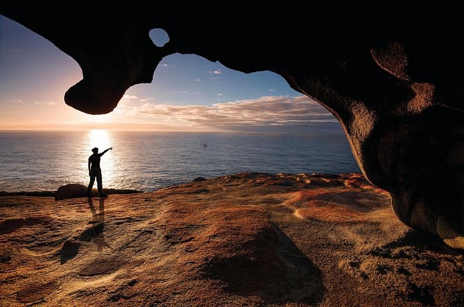 A walker silhouetted under a wind-hollowed granite boulder at sunset over the sea